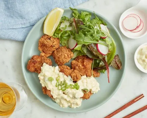 Crispy Japanese chicken karaage topped with creamy tartar sauce and chopped green onions, served on a light green plate with a side salad, radishes, a lemon wedge, and chopsticks.