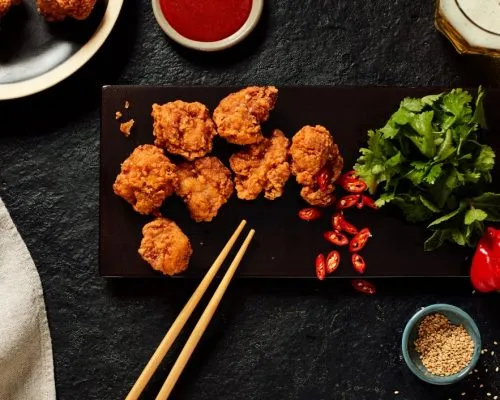Crispy Japanese chicken karaage served on a black platter with sliced red chilies, fresh coriander, sesame seeds, and dipping sauce.