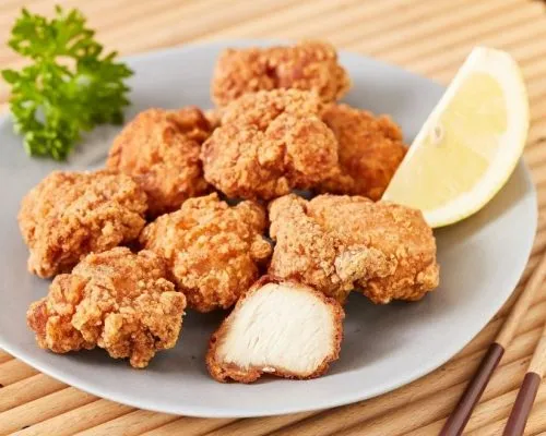 A plate of golden-brown Japanese chicken breast karaage with one piece cut open to show the inside, served on a grey plate with a lemon wedge and parsley garnish on a bamboo mat.