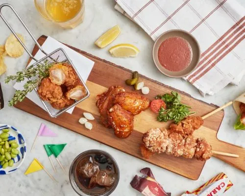 A Japanese chicken karaage party platter featured on a wooden serving board alongside chicken wings, skewers, a mini frying basket of karaage, edamame, lemon wedges, dipping sauce, and drinks.