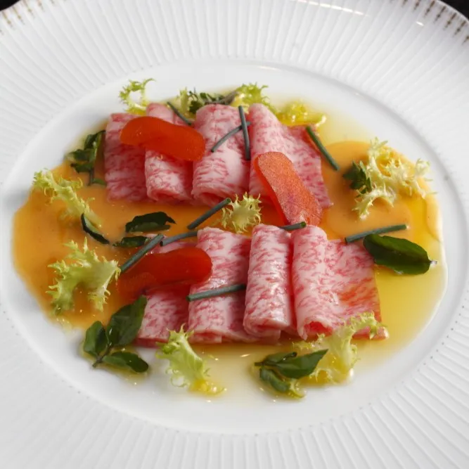 Thinly sliced, melt-in-the-mouth Miyazaki Wagyu beef carpaccio elegantly arranged with a vibrant orange citrus dressing and fresh microgreens on a white fluted plate.